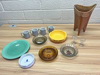 Full lot on wooden table showing ashtrays, mugs, Fiestaware plate and other glassware including a copper vase.