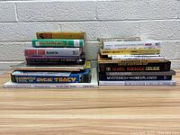 Two stacks of assorted books on tabletop in front of white brick wall, showing spines and covers with visible titles.