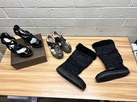 Lot overview showing all three pairs of shoes on a wooden table against a concrete wall backdrop: Bebe black heels on a shoe box, brown and beige Ecco shoes, and mid-calf Skechers black boots with rhinestones and fur lining.