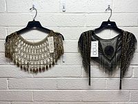 Two women's beaded shawls hanging on black hangers against a white brick wall. One gold with tassel beads, one black with gold bead accents and fringe.