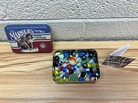 Lot showing the metal tin, glass marbles inside the tin, and instruction booklet all displayed against a wall.