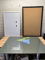 Photo of magnetic dry erase whiteboard with two round magnets and black framed corkboard displayed on tabletop with glass chalkboard panel and product label visible.