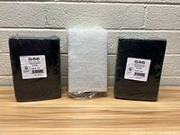 Lot of three nylon scrub pads: two black heavy duty and one white pad. Shown against a brick background.