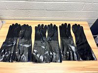 Three pairs of black industrial gloves laid side by side on a wooden surface, showing full length and shiny surface.