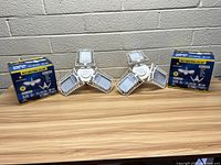 Pair of new multi-directional LED security lights displayed with their packaging boxes on a wooden surface against a brick wall.