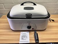 Front view of Rival 18 quart electric roaster oven on wooden table, showing front temperature control dial, on/off indicator light, removable lid with handle, detachable roasting rack handle, and owner's manual booklet.