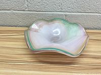 Glass dish showing side profile with wavy edges and multi-color swirl pattern including green, pink, and white hues on a wooden background.