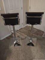 Pair of black bar stools with chrome bases shown from back side against a white paneled door and carpet floor