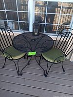 Round black metal mesh bistro table with two matching chairs having green cushions, placed on a wooden deck.
