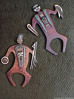 Two tribal-style decorative wall art figures displayed on a green textured fabric. Both figures are stylized with geometric patterns and hold different objects. The smaller figure has feathers attached to its head.