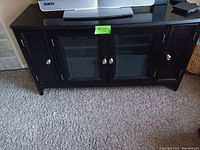 Full view of black wooden cabinet showing the front with two central glass doors and two side wooden doors, placed on carpeted floor with a TV on top.