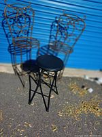 Two metal iron bar stools with aged rust finish and decorative scrollwork backs and seats, accompanied by one small black folding stool with round padded seat and metal frame, photographed on asphalt with blue industrial door background.