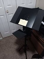 Full view of black metal adjustable music stand with side shelf, standing on tripod base indoors near closet doors.