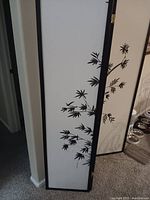 Three-panel room divider standing upright near a corner, showing black frame and white panels with black bamboo leaf design.