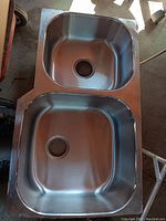 Top view photo of a stainless steel double basin sink showing two large rectangular basins with drain holes, placed on a flat surface.