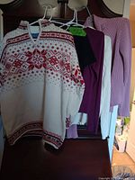 Four women's sweaters/shirts hanging on hangers, showing details of color and knit patterns.