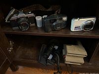 Overview of all cameras and binoculars on wooden shelf, includes film rolls