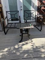 Two dark metal patio chairs with geometric lattice backs and armrests, positioned around a small matching metal table with slatted top on wooden deck in front of glass doors.