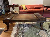 Full side view of rectangular wooden coffee table with curved legs and metal claw feet sitting on a patterned carpet by a red couch and a round side table.