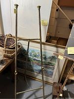 Full view of brass easel positioned in front of a framed painting, showing pitting and overall condition.