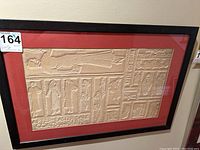 Front-facing view showing entire framed relief art with beige 3D figures and text on red backing inside black frame.