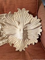 Off-white ceramic round sun face garden decoration with leaf rays, sitting on brown textured fabric couch.