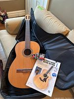 Wooden classical acoustic guitar resting inside a black soft gig bag, alongside a guitar instruction manual and a clip-on tuner on the couch.