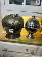 Photo of two liqueur sets on a wooden counter, one designed like a medieval armor chest piece and the other a spherical stainless steel decanter with lid and handles.