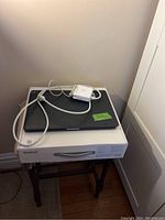 Black MacBook closed with power adapter on top, placed on white box on small black table.