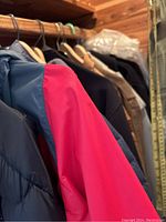 Close-up of black and red color-block puffer jacket sleeve with wooden hanger and other jackets behind