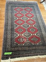 Full view of rectangular oriental rug showing red hexagonal motifs on a black background, surrounded by intricate cream and gray borders, laid on wooden floor.