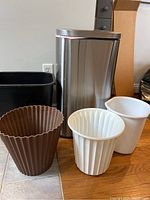 Five garbage cans including one large stainless steel step-on kitchen trash can and four smaller plastic garbage cans of various colors and styles on floor.