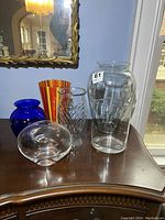 Overview of all six vases on wooden surface including tall clear vase, cobalt blue vase, small flat-bottom clear vase, textured small clear vase, striped orange-red Kosta Boda vase, and oval clear vase.