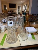 Wide view of assorted glass pitchers and vases including clear glass pitcher with patterned sides, blue and orange glass vases with toppers, and milk glass pedestal bowl.