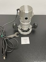 Front view of Craftsman router showing metal casing, 1/4 inch collet, built-in worklight, black ergonomic handles, attached power cord, and metal wrench included.