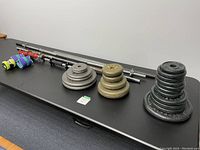 Photo of various weights, bars, and ankle weights arranged on a black table, showing their size and condition.