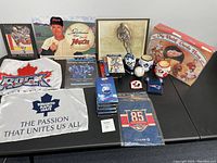 Various sports memorabilia items arranged on table including Toronto Maple Leafs banners, DVDs, VHS tapes, collectible mugs, Gordie Howe cereal boxes, and framed cycling artwork.