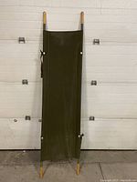 Photo of vintage collapsible field hospital stretcher standing upright showing wooden poles and green fabric.