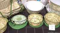Overview of multiple melamine serving bowls, plates, and bakers arranged with black wrought iron holders and green metal trivets, showing coordinated green and beige floral patterns.