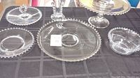 Six clear glass Candlewick pieces on black tablecloth. Pieces include plates, bowl, cake plate, fan vase and handled server all with beaded edges.