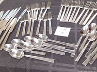 Photo showing knives, dessert forks, dinner forks, teaspoons, and serving spoons laid out on a dark surface.