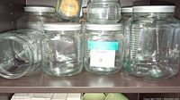 Eight glass storage jars on dark wood shelf. Varied sizes, clear glass, white lids. One jar filled with wooden clothespins.