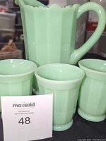 Jadeite glass pitcher and 4 cups arranged with MaxSold card showing lot number
