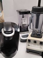 Photo showing Keurig coffee maker, newer Ninja blender, and older model Vita-Mix blender on a white surface against plain background.