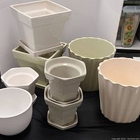 Overall view of all 8 ceramic plant pots showing various shapes, sizes, and colors including cream, pale green, and white with some matching saucers.
