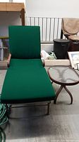 Photo showing full view of the metal chaise lounge with green cushion and its adjustable backrest, along with the matching side table with glass surface beside it.