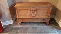 Front view of the wooden cedar chest showing carved floral design on front, lower drawer with metal handles, turned wood spindles on corners, and bun feet.
