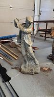 Front view of weathered garden figure of a golfer mid-swing wearing traditional golf attire, standing on a textured base.