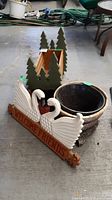 Photo of garden pot, welcome sign with swans, and wooden accessory box with tree cut-outs on a concrete garage floor.