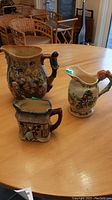 Three assorted ceramic jugs on wood table, two Royal Doulton and one Crown Devon music box jug, showing general size and decoration